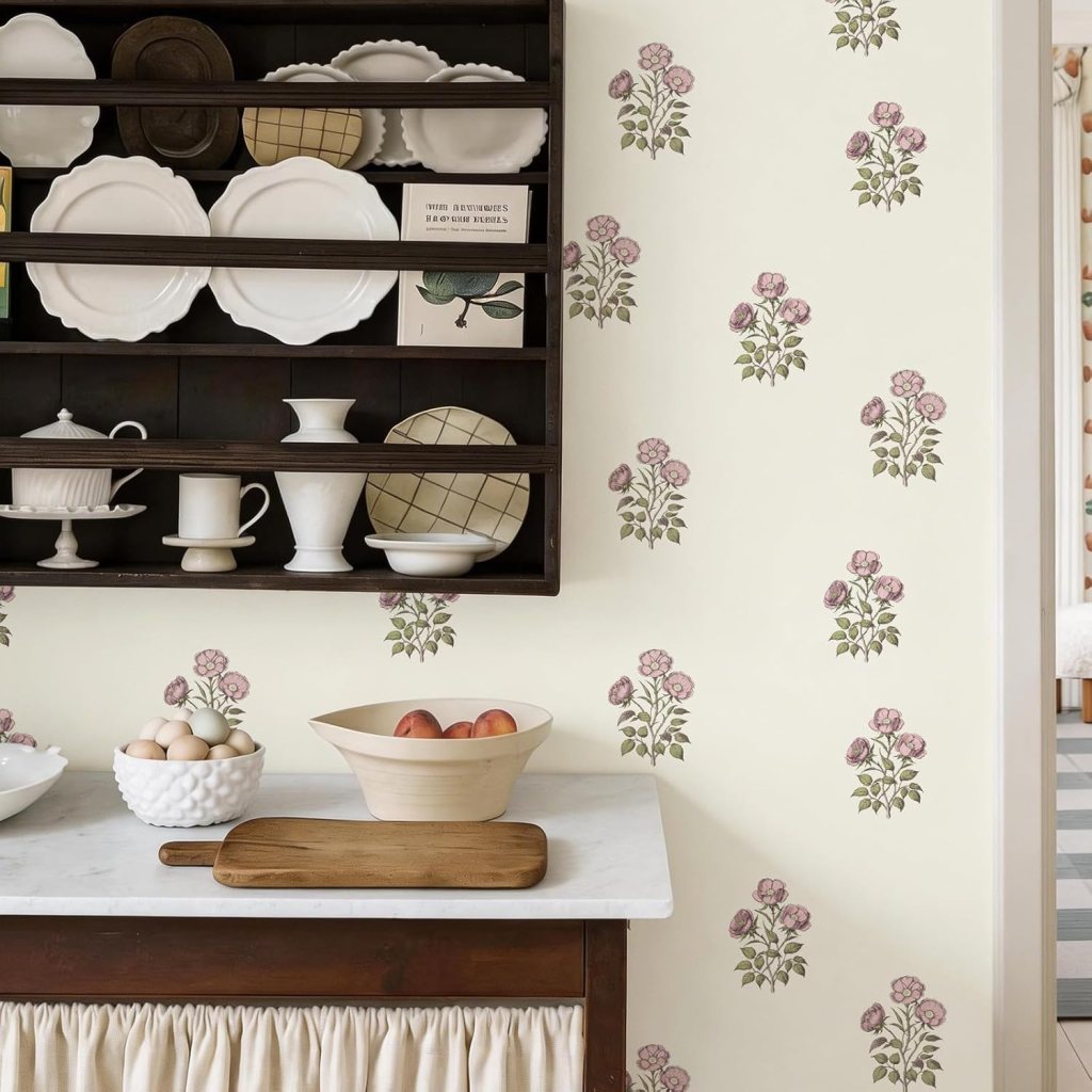 A cozy kitchen scene featuring a wooden shelf with white dishes and a book against a floral wallpaper. A marble countertop displays a bowl of apples and eggs, with a wooden cutting board nearby.