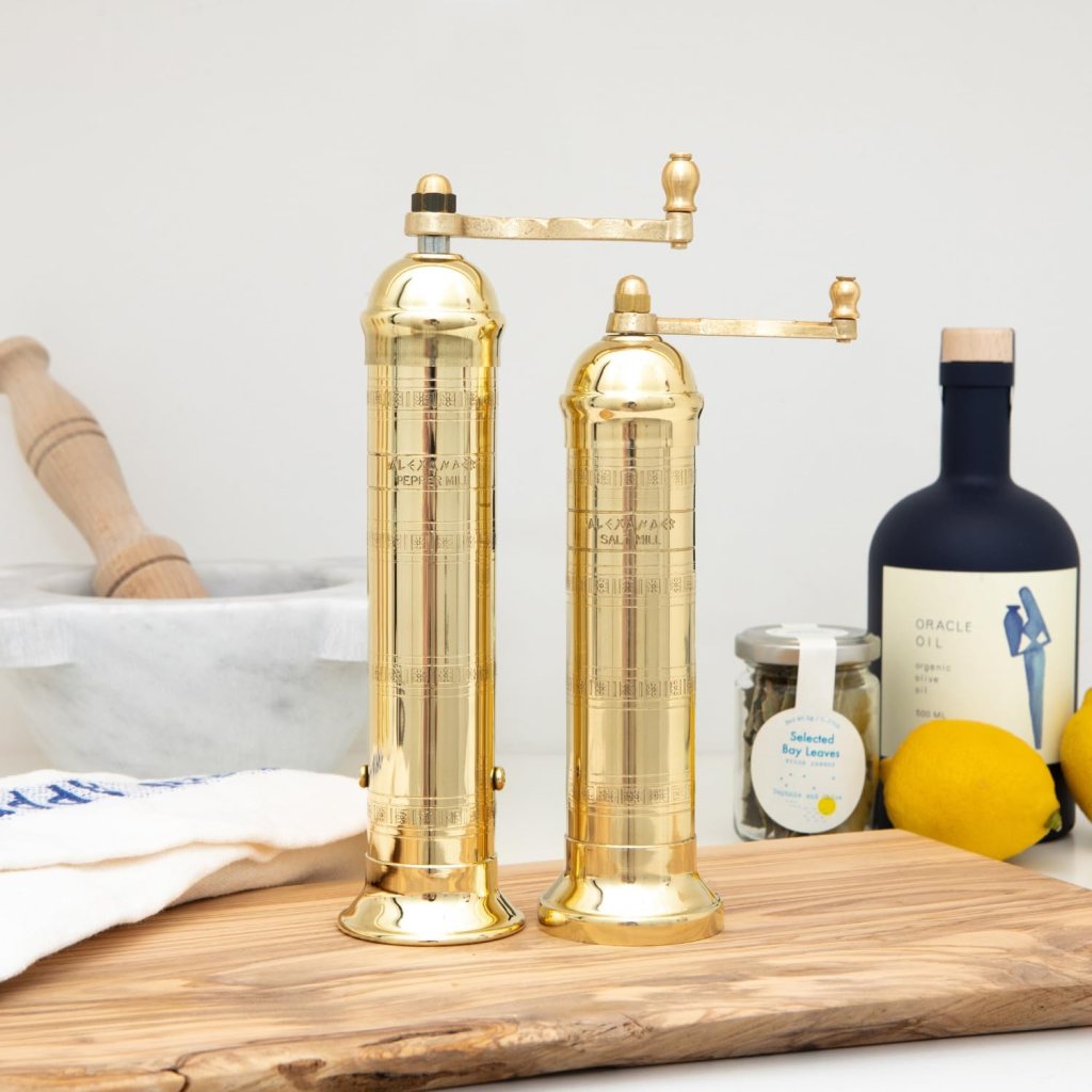 Two brass pepper and salt mills placed on a wooden cutting board, with a marble mortar and pestle, a bottle of olive oil, a jar of bay leaves, and a couple of lemons in the background.
