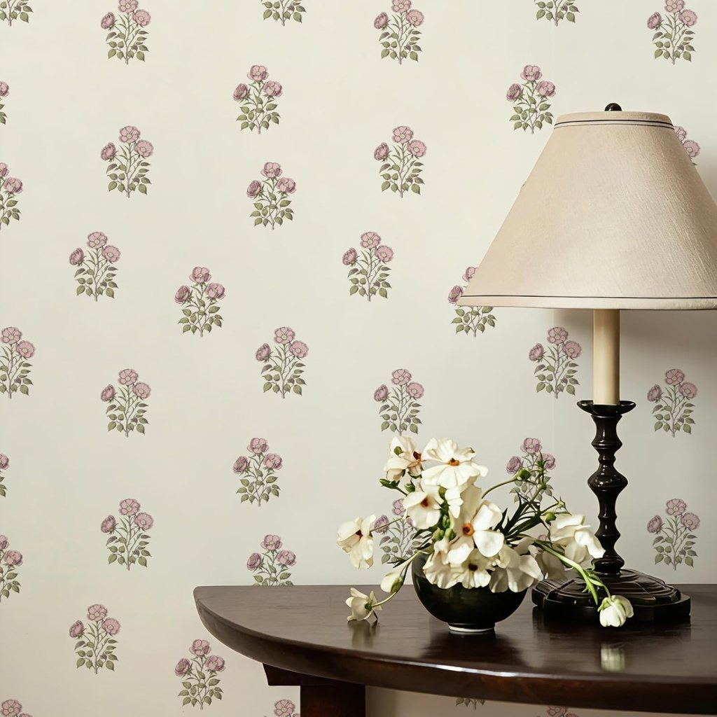 A cozy interior scene featuring a wooden table with a black vase holding white flowers, beside a lamp with a beige shade, against a floral wallpaper background.