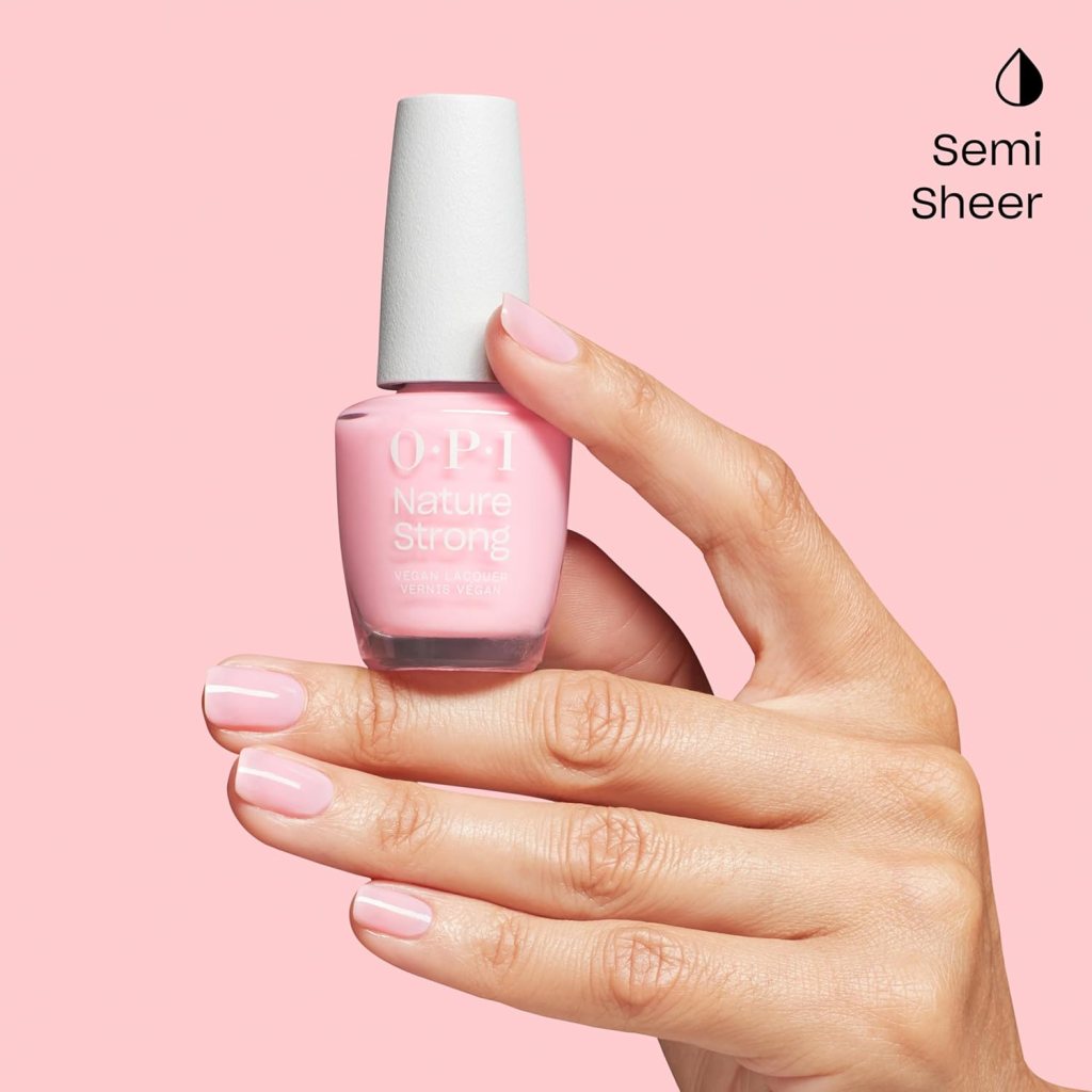 A hand holding a bottle of OPI Nature Strong semi-sheer pink nail polish, with well-manicured nails painted in a matching shade, against a pink background.