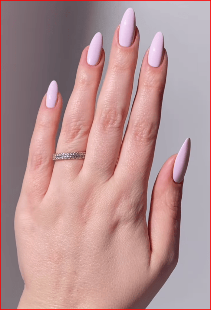 A close-up of a hand with long, almond-shaped nails painted in a light pink shade, wearing a silver band ring.