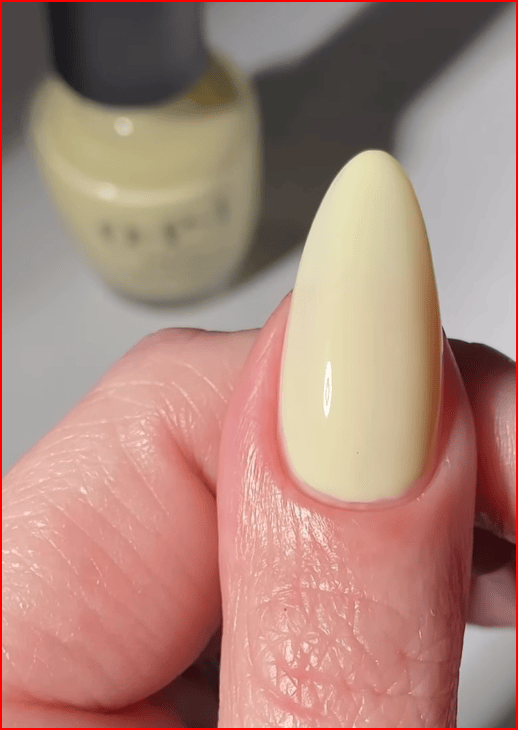 A close-up of a hand holding a sharp, pointed nail painted in a light yellow nail polish, with a bottle of the same nail polish in the background.